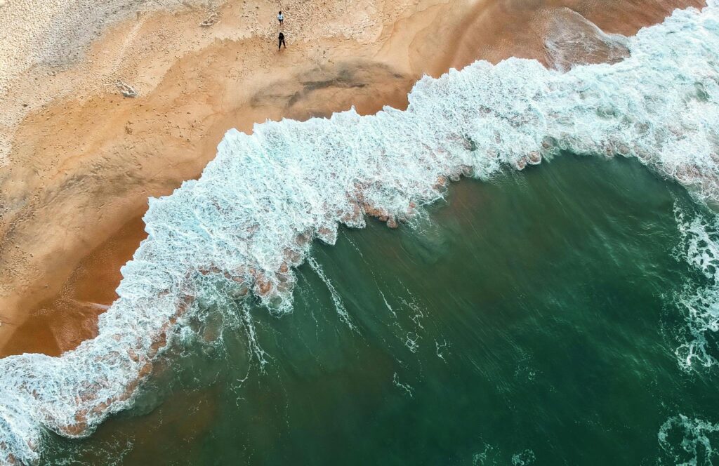 Aerial shot of ocean waves crashing onto a sandy beach with two people in the distance.